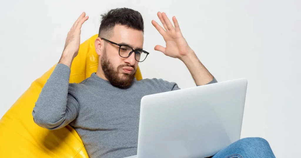 A man with glasses sits on a yellow bean bag, raising his hands in frustration while looking at a laptop screen demonstrating a bad ad copy.